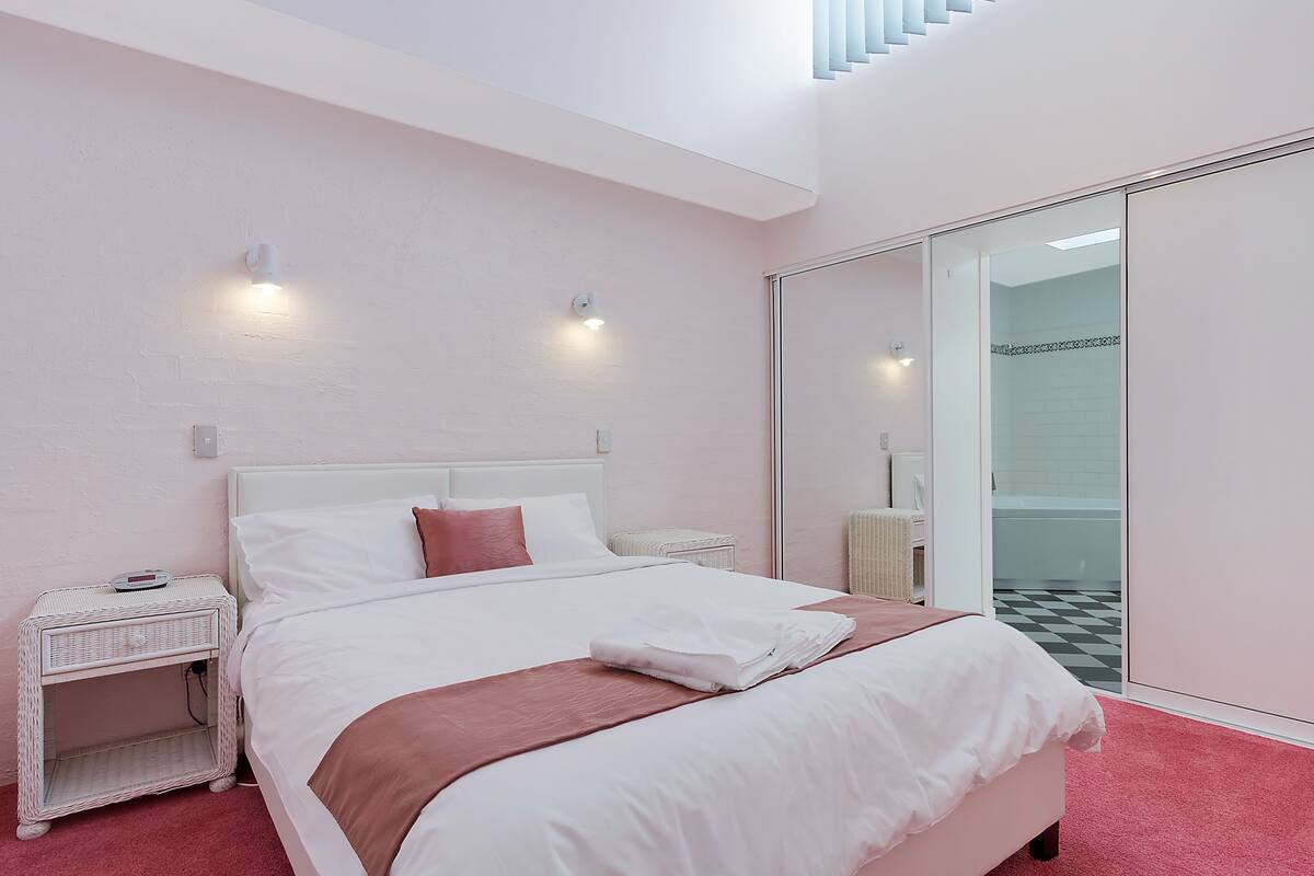 A bedroom with a white bed, pink accents, a white nightstand, and a glass door leading to a bathroom.