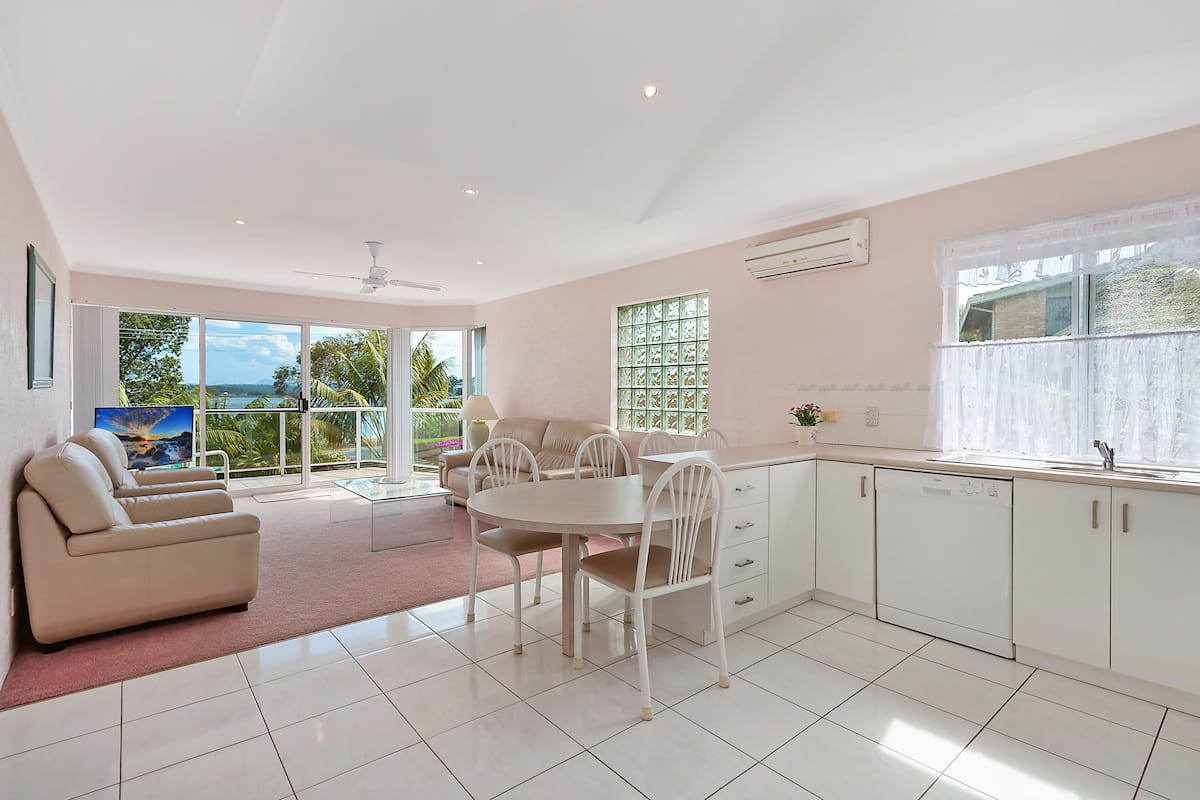 A modern kitchen with white cabinets, a dining table, and chairs. There is a large window with a view of greenery and a building.