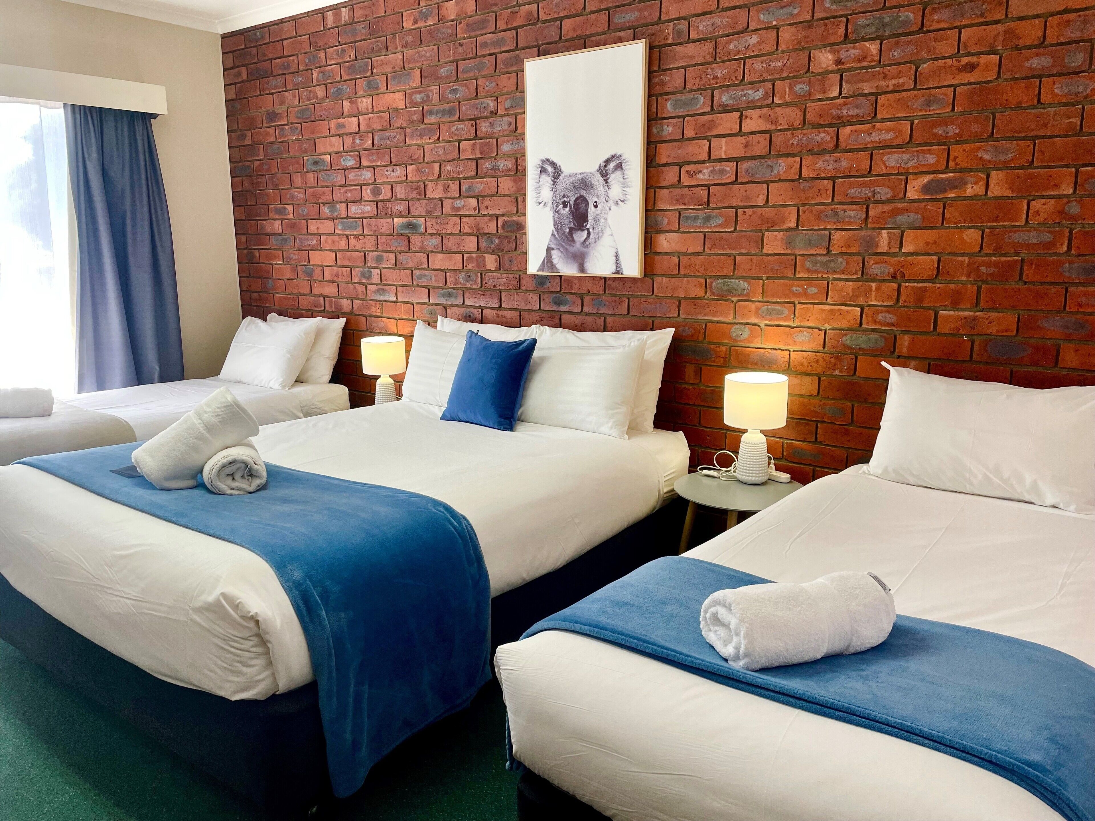 Habitación de hotel con dos camas, una pared de ladrillo, un cuadro de koala y una ventana con cortinas.