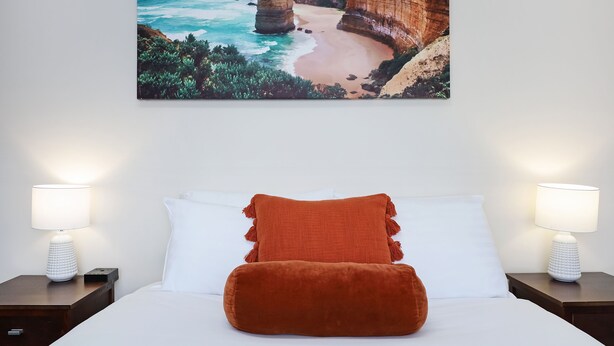 A bedroom with a bed, two nightstands, a white lamp, a brown pillow, and a coastal landscape painting on the wall. - City Heart Motel (Warrnambool)