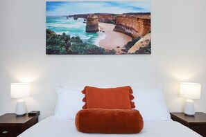 A bedroom with a bed, two nightstands, a white lamp, a brown pillow, and a coastal landscape painting on the wall. - City Heart Motel (Warrnambool)