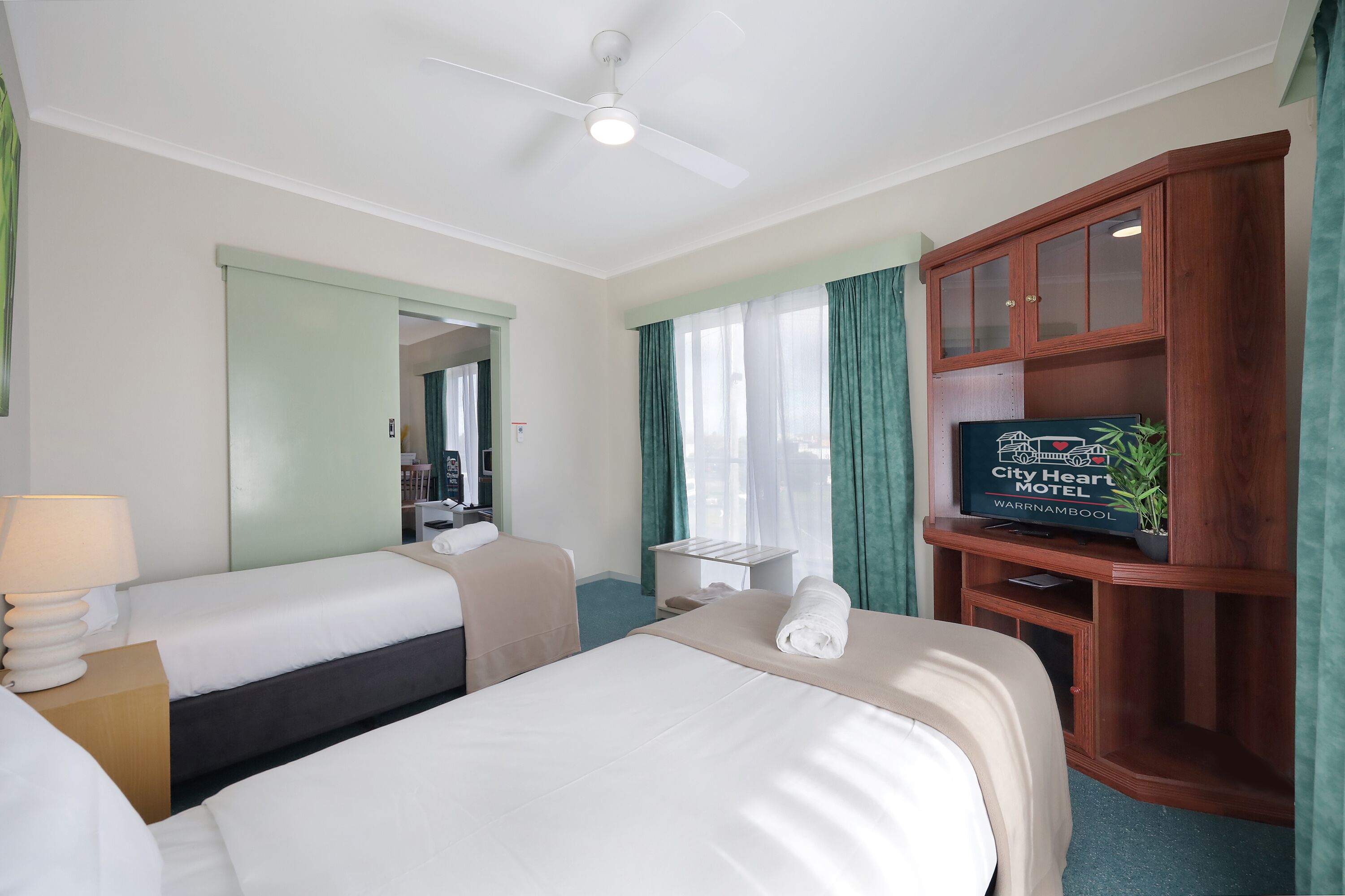 Ein Hotelzimmer mit zwei Betten, einem Deckenventilator, einem Fernseher, einem Holzkabinett und einem Fenster mit Vorhängen.
