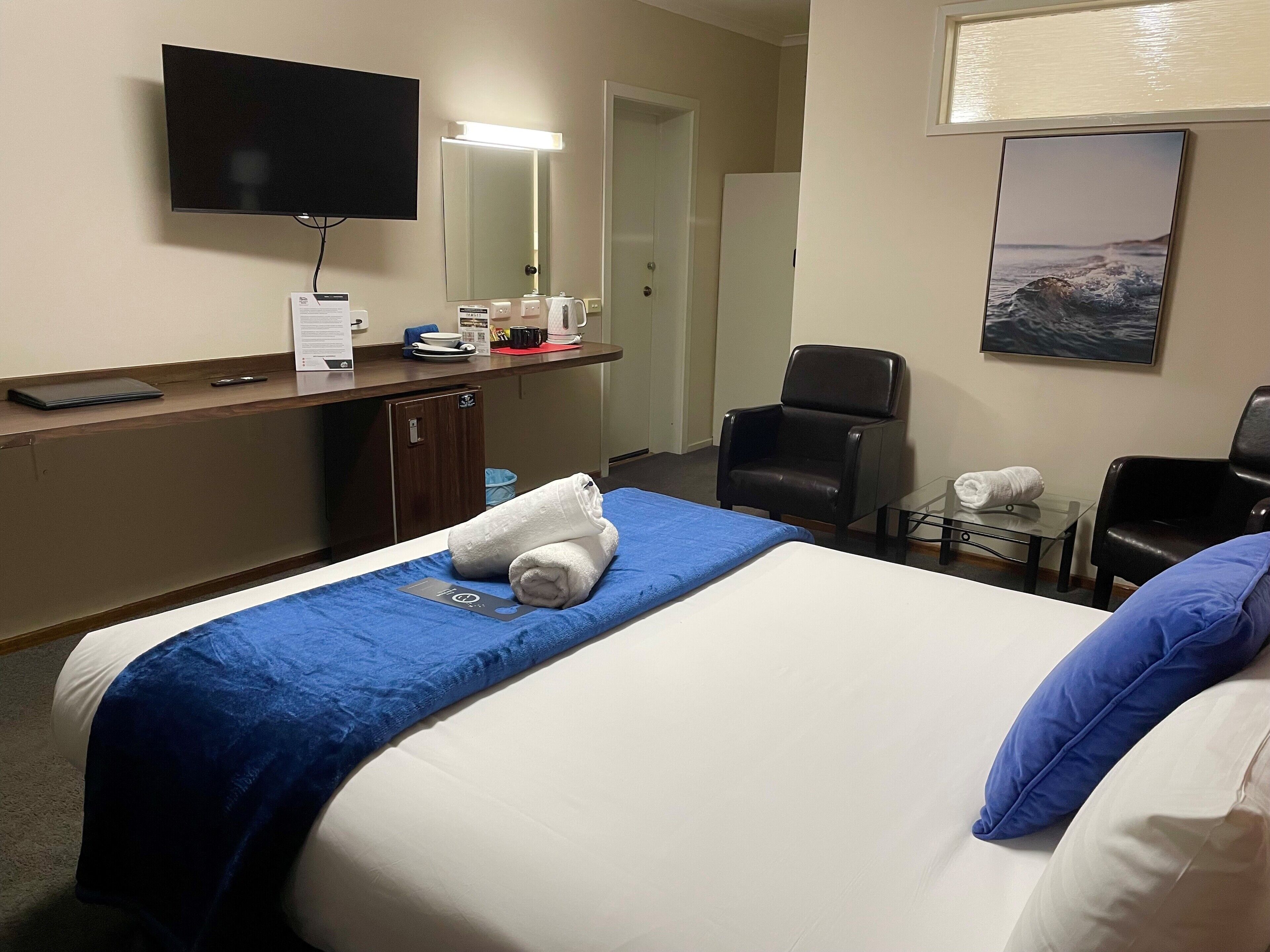 Ett hotellrum med en säng, ett skrivbord med en platt-TV, en stol och ett litet bord med en handduk.
