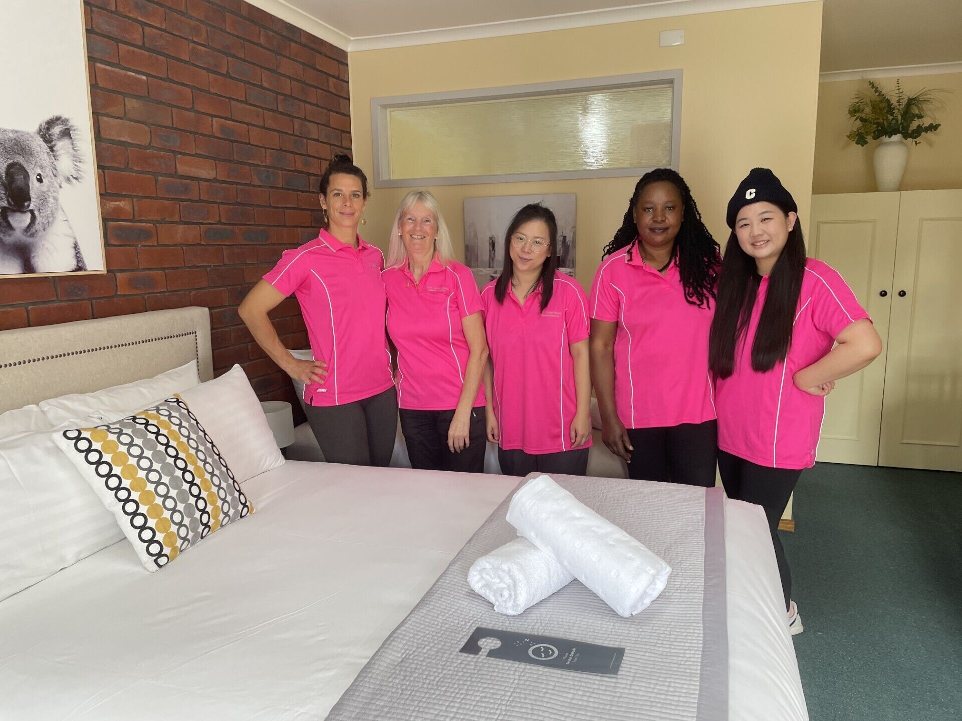 A group of people in pink shirts standing in a hotel room with a bed, pillows, and a framed picture on the wall.