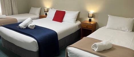 Habitación de hotel con dos camas, cada una con sábanas blancas y una manta azul en una cama. Hay un almohadón rojo en la cama central. La habitación tiene una mesita de noche de madera con una lámpara a cada lado de las camas.