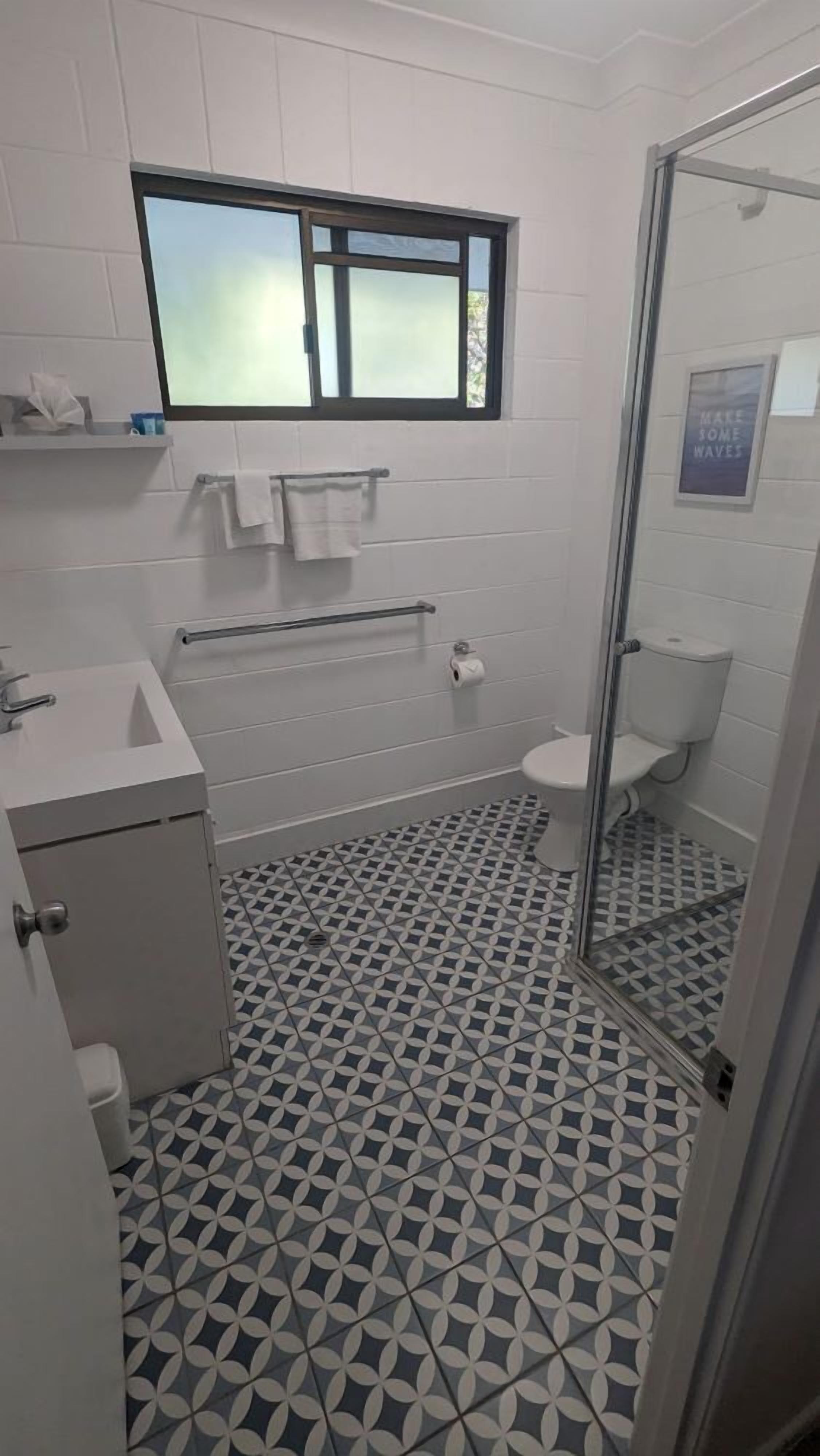 A bathroom with a geometric tile floor, a toilet, a sink, a glass shower enclosure, and a window above the sink.