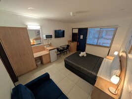 Habitación compacta con cama, escritorio con computadora, refrigerador pequeño, sofá y ventana con persianas.