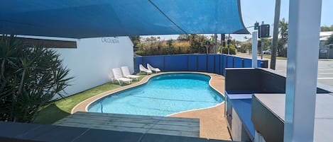 A swimming pool with lounge chairs, surrounded by a blue fence and a shaded area.