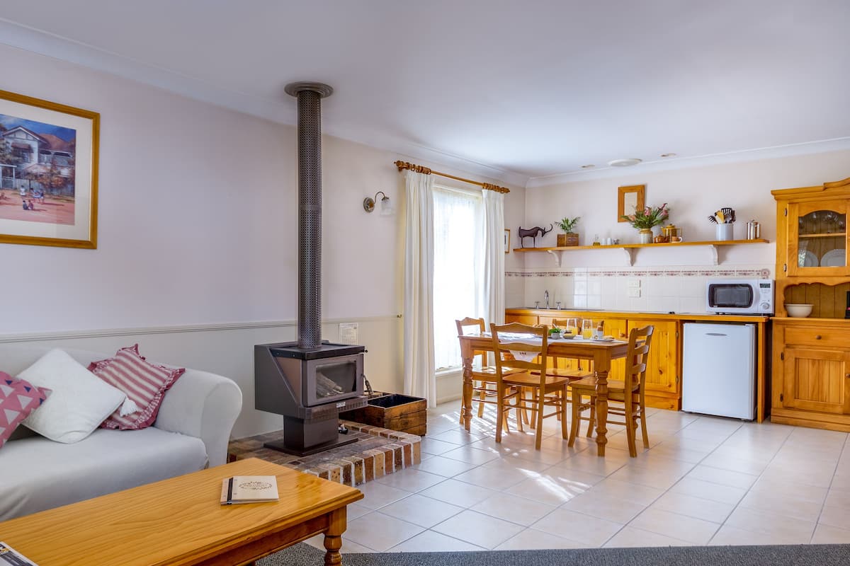 Una sala de estar con un sofá, una mesa de centro y una chimenea. Hay una zona de cocina con microondas, refrigerador y armarios de madera. Un cuadro enmarco cuelga de la pared.