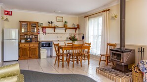 A cozy kitchen with wooden cabinets, a dining table, chairs, a microwave, a refrigerator, a fireplace, and a fire blanket sign on the wall. - Honeysuckle Cottages Stanthorpe (Applethorpe)