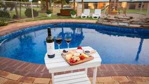 A poolside setting with a table set for a meal, including a bottle of wine, glasses, and a cheese board.