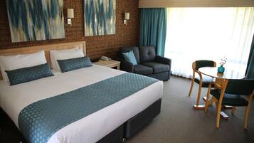 A hotel room with a large bed, a grey armchair, a wooden table, and two wall-mounted lamps.