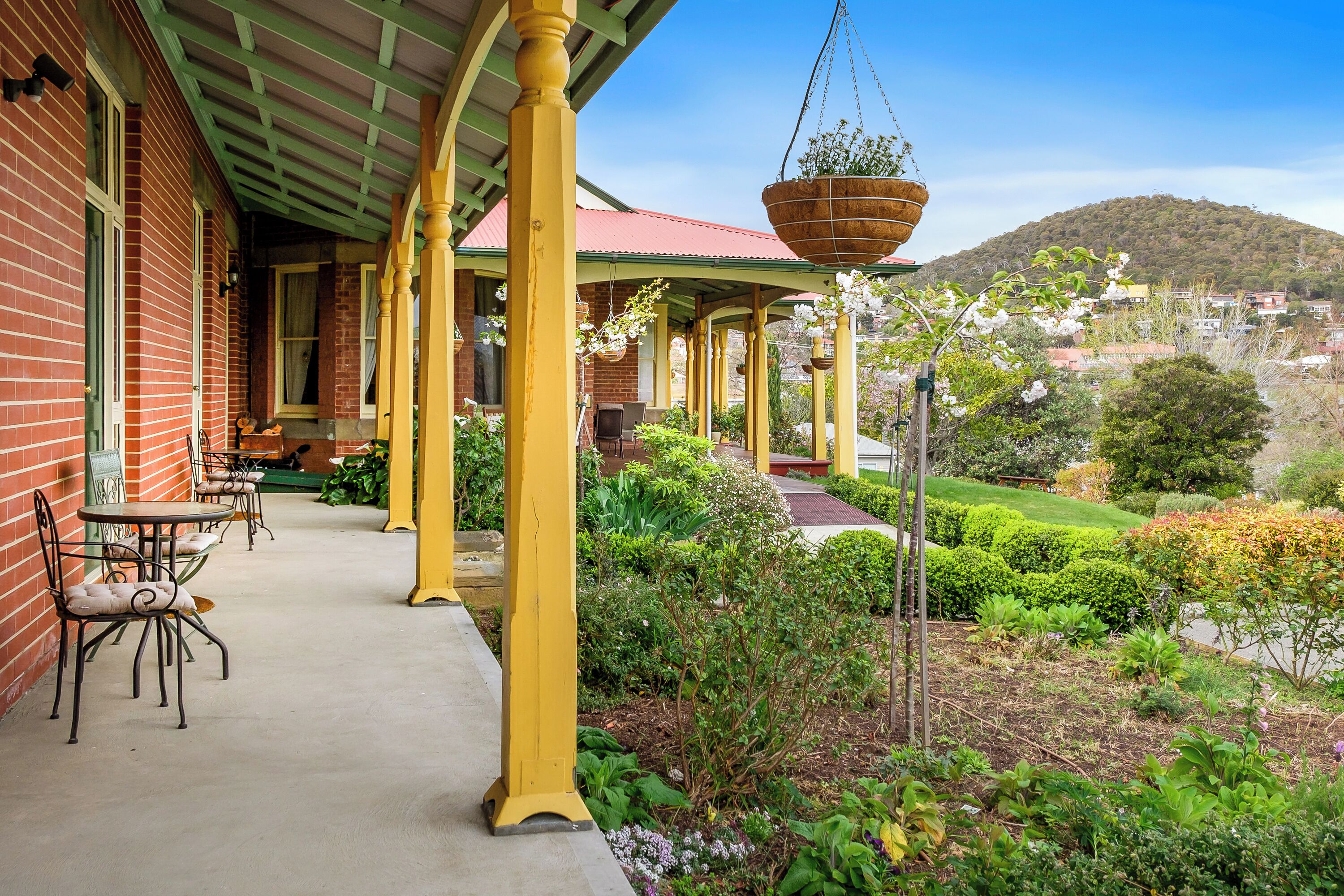 A covered outdoor area with a brick building, yellow pillars, and a garden with various plants and flowers.