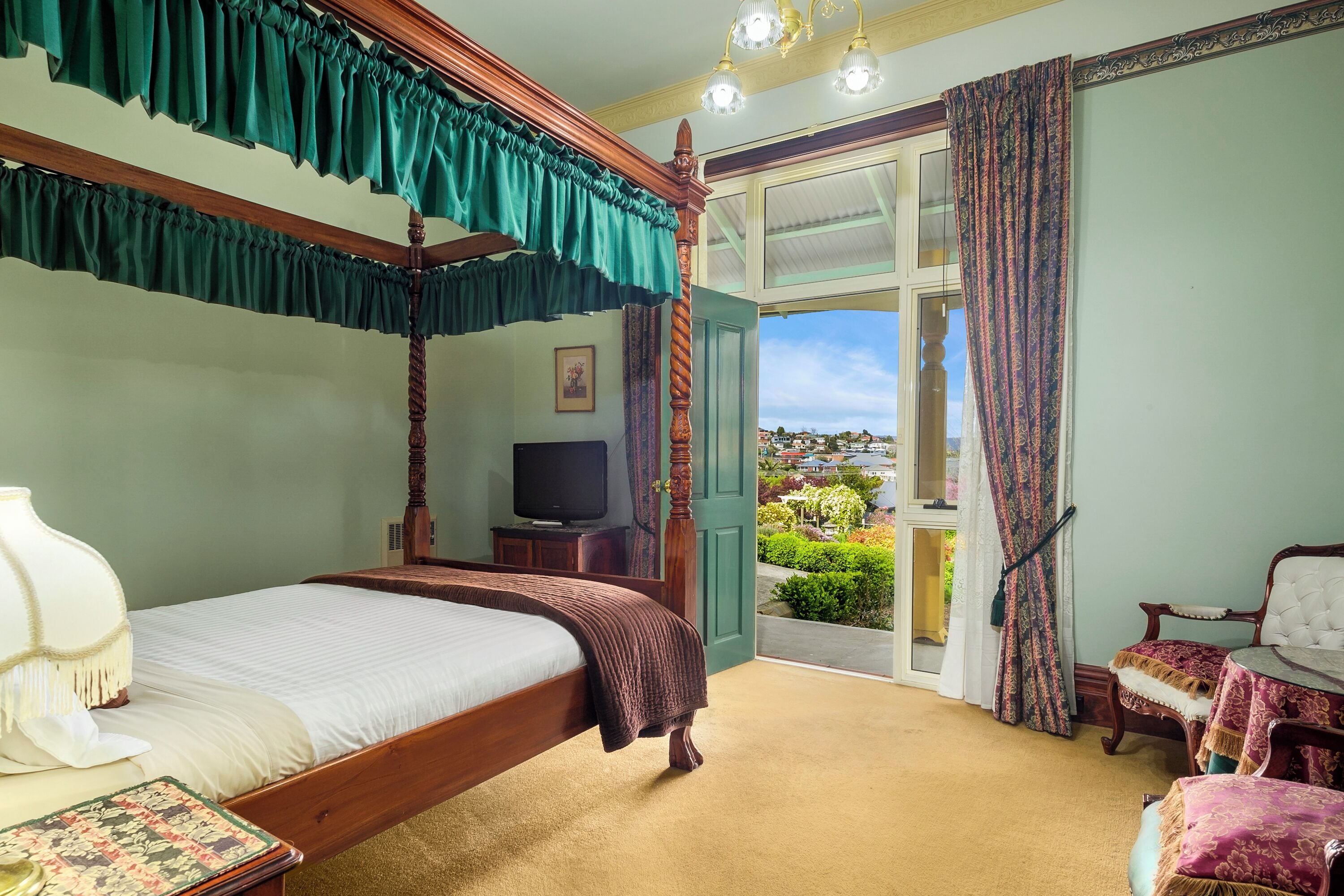 A four-poster bed with a television on a stand, a chair, and a view of the outside.