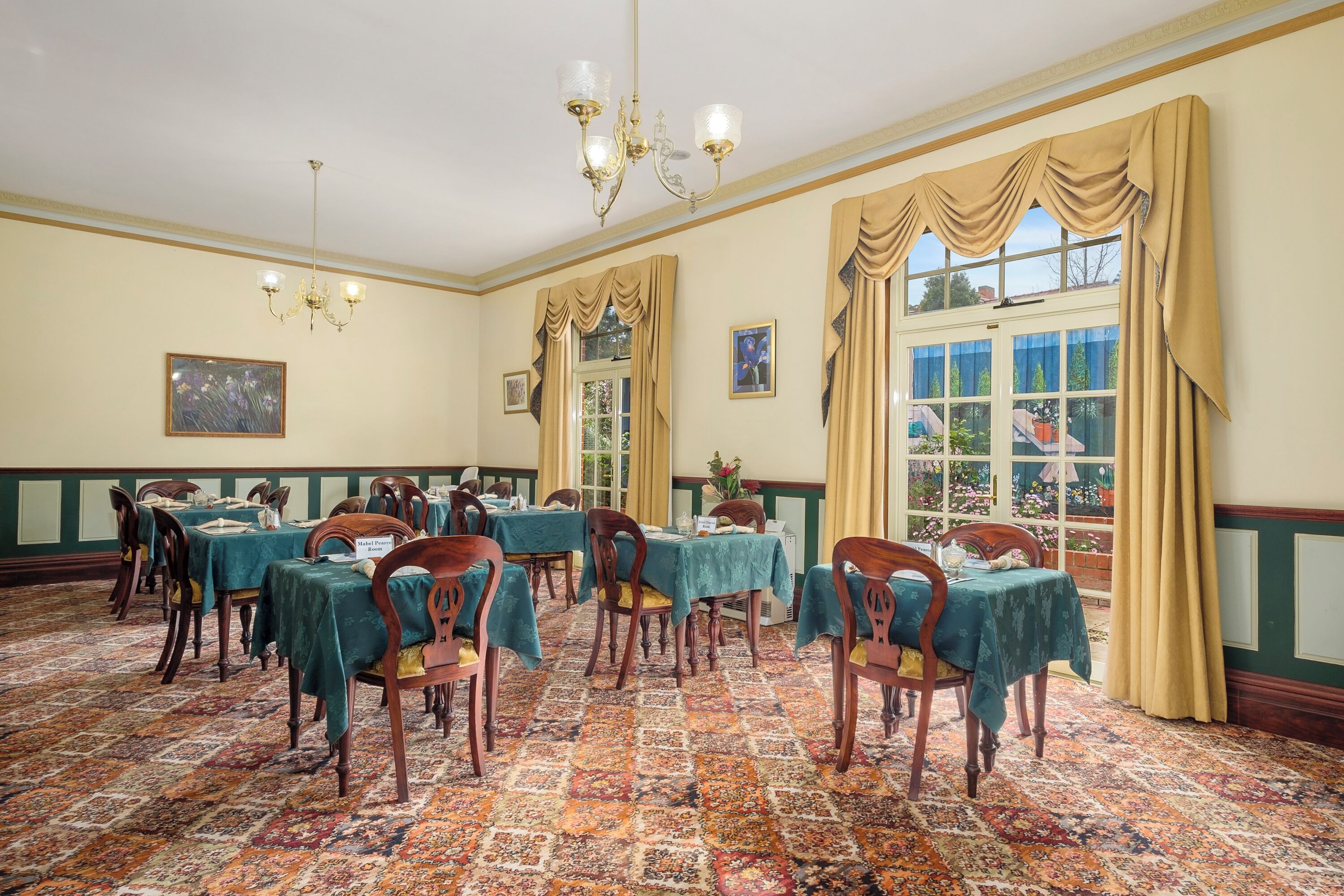 A dining room with tables set for a meal, a chandelier, and large windows with curtains.