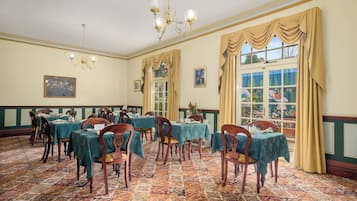 A dining room with tables set for a meal, a chandelier, and large windows with curtains.