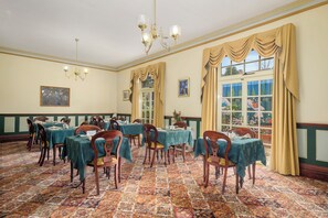 A dining room with tables set for a meal, a chandelier, and large windows with curtains. - Orana House (Lindisfarne)