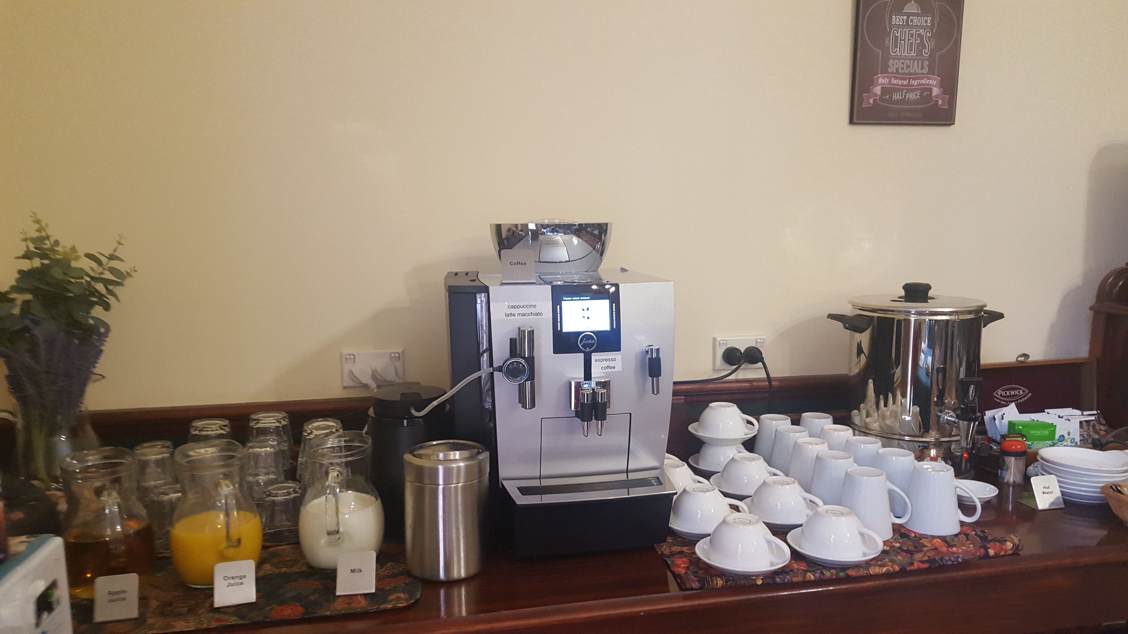 A coffee station with a coffee machine, milk, and juice dispensers, and a selection of white cups and saucers.