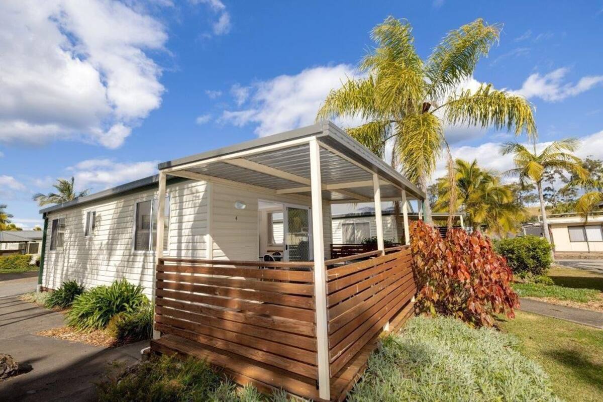 A single-story mobile home with a covered porch, surrounded by tropical vegetation and palm trees.