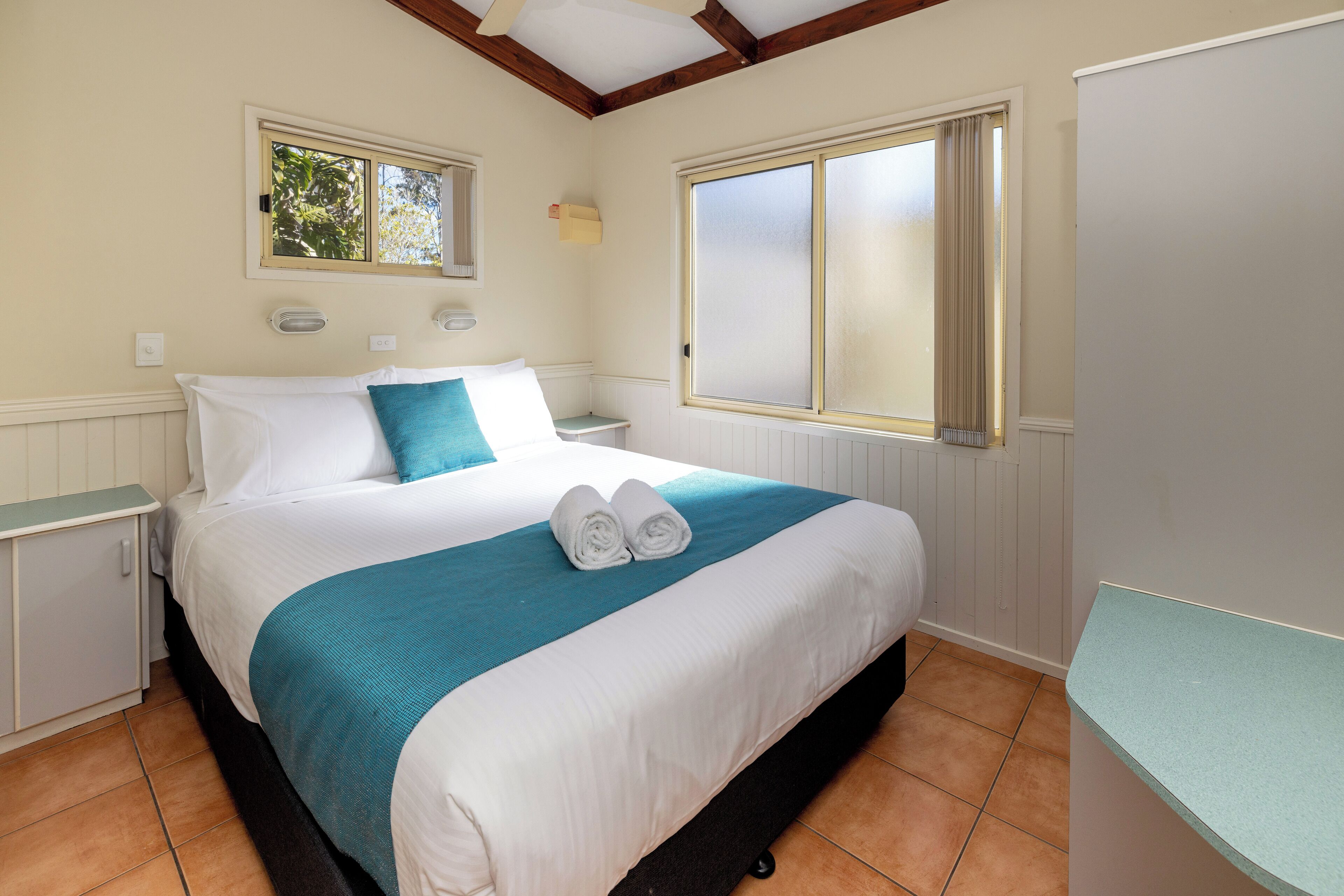 A neatly made bed with white and teal bedding, two bedside tables, and a window with curtains.