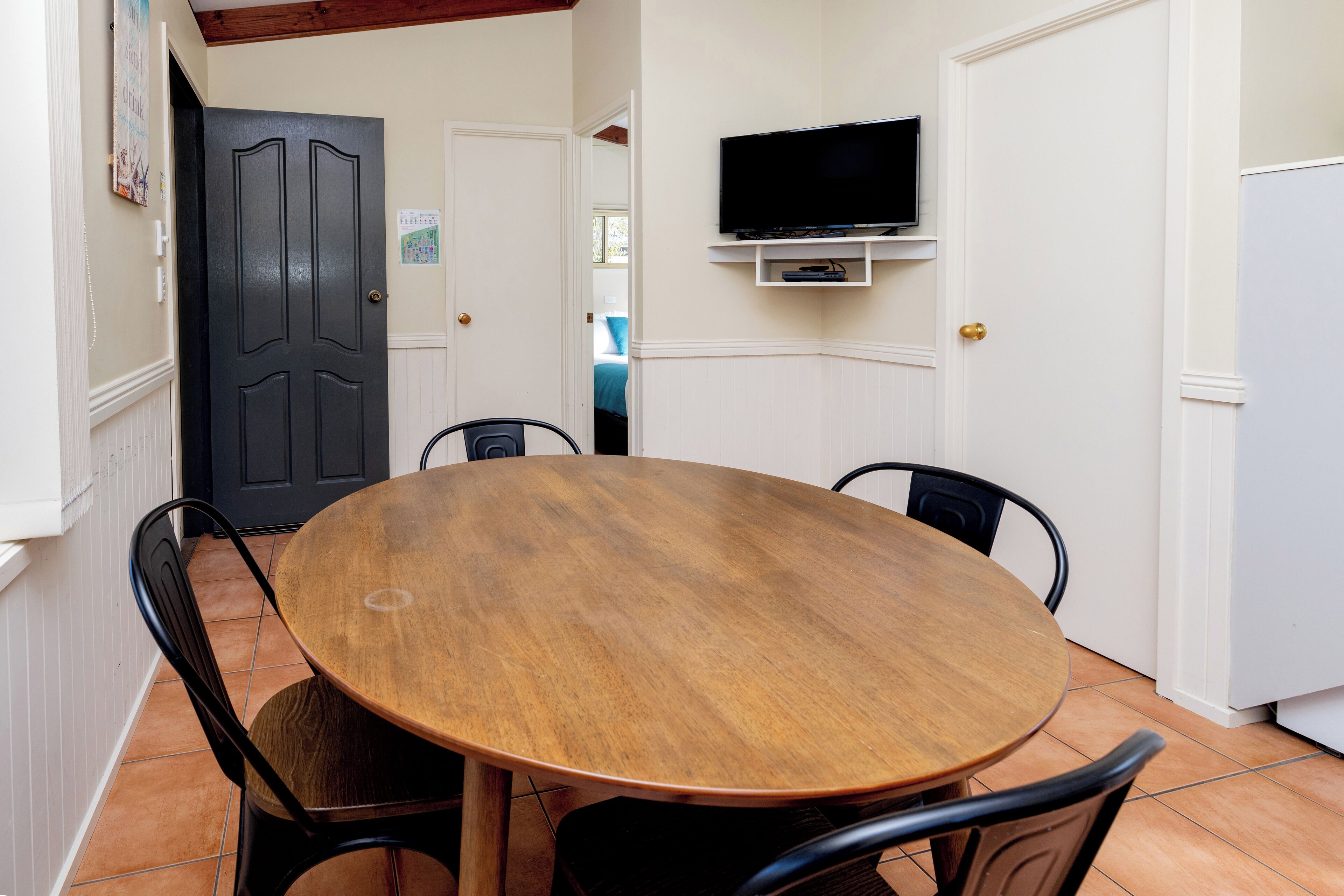 A dining area with a round wooden table and four chairs, a television mounted on the wall, and a door leading to another room.