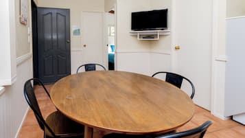 A dining area with a round wooden table and four chairs, a television mounted on the wall, and a door leading to another room.