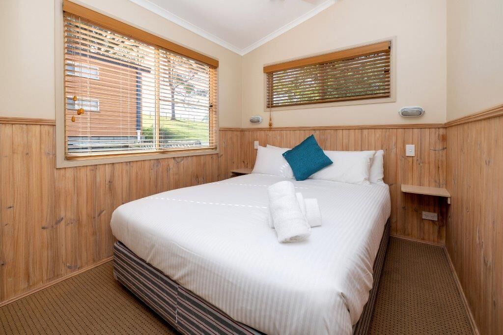 A neatly made bed with white linens and a blue pillow, a wooden headboard, and a window with blinds.