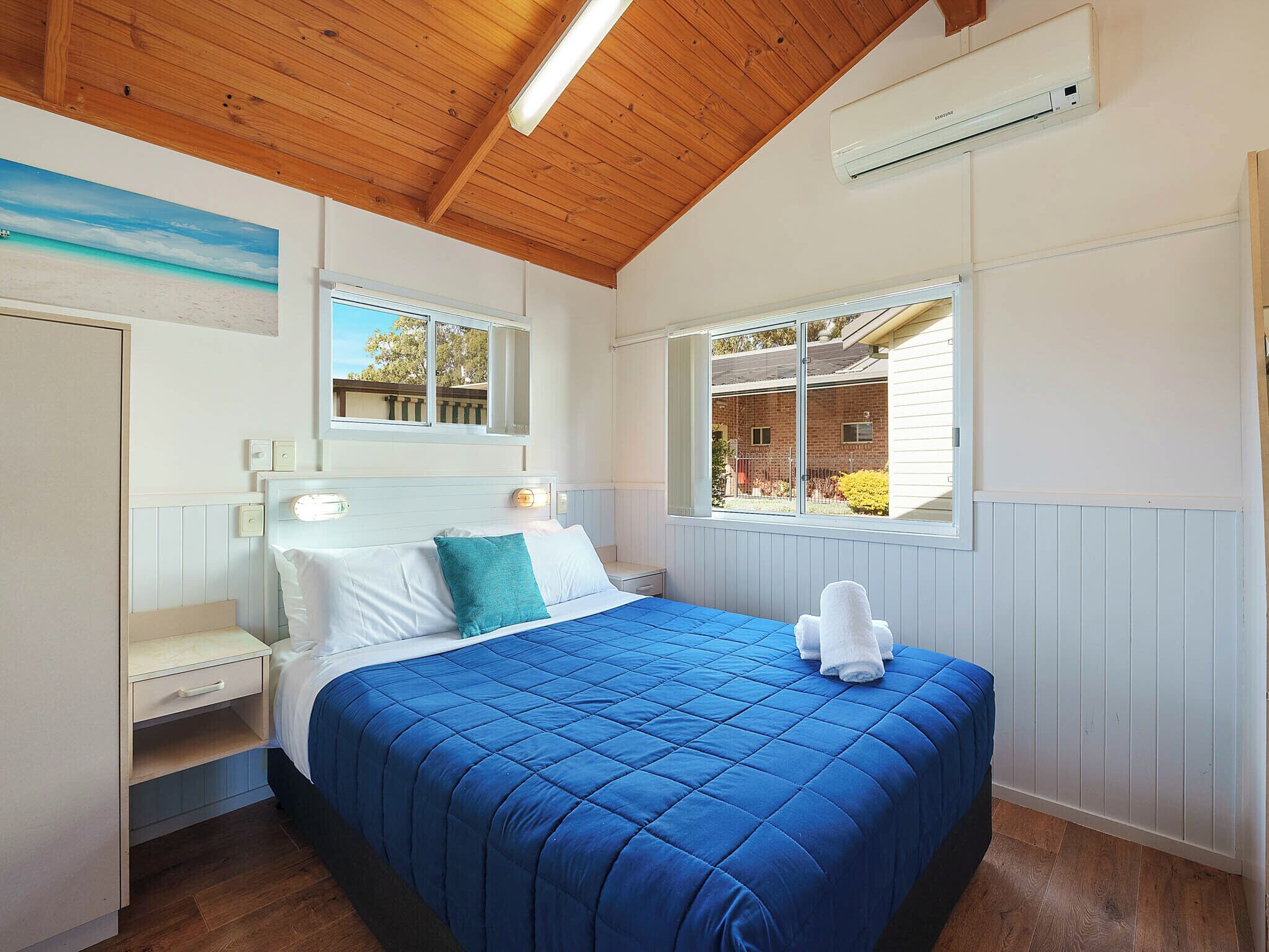 A bedroom with a wooden ceiling, a bed with a blue quilt, a nightstand, a bedside table, a window with a view of the outside, and an air conditioning unit.