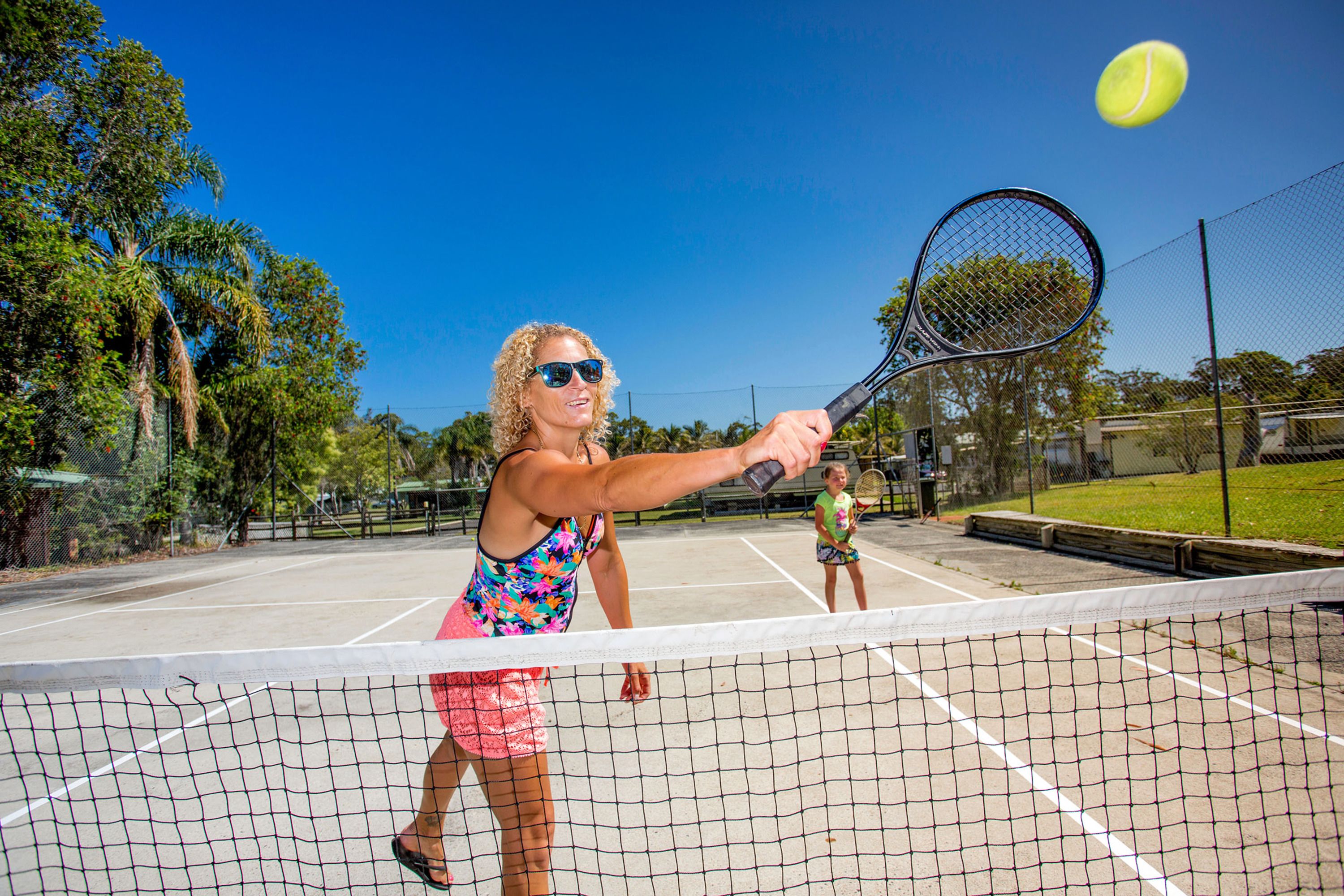 A tennis court with a net in the foreground, a player in the background, and a tennis ball in mid-air.