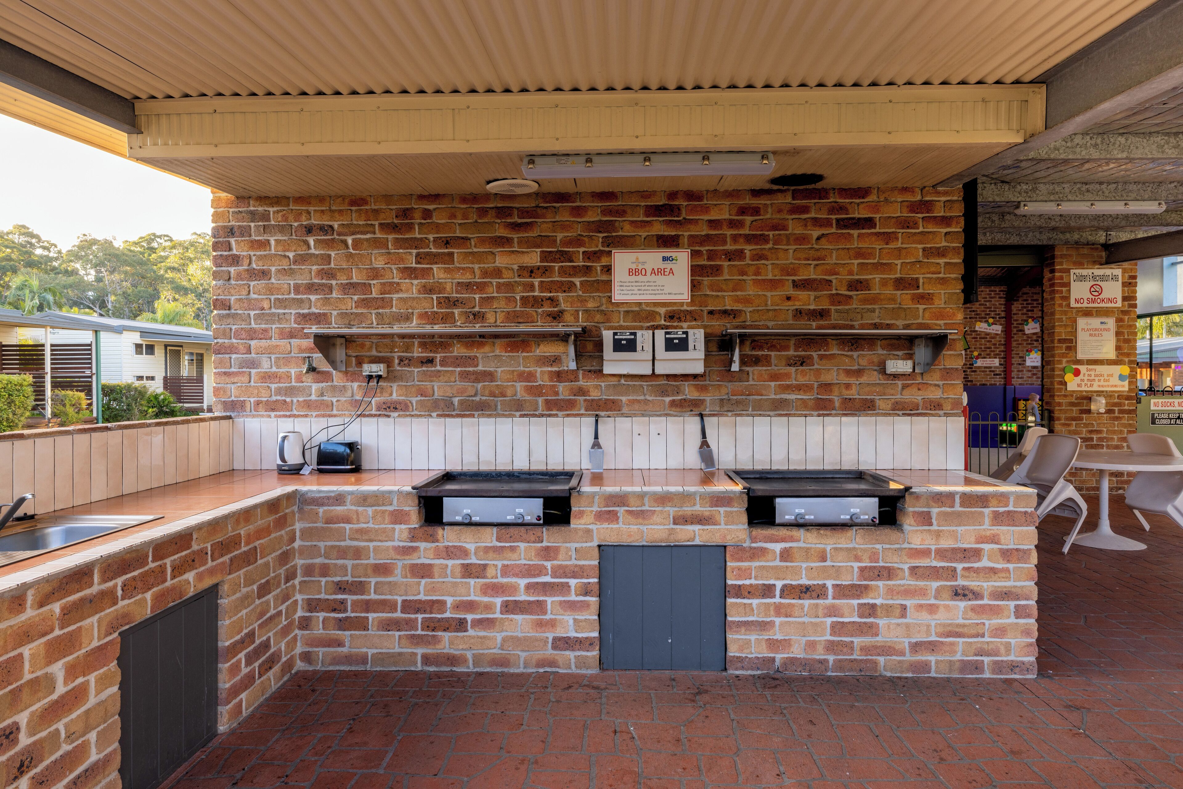 An outdoor brick barbecue area with built-in grills, a sink, and a children's recreation area.