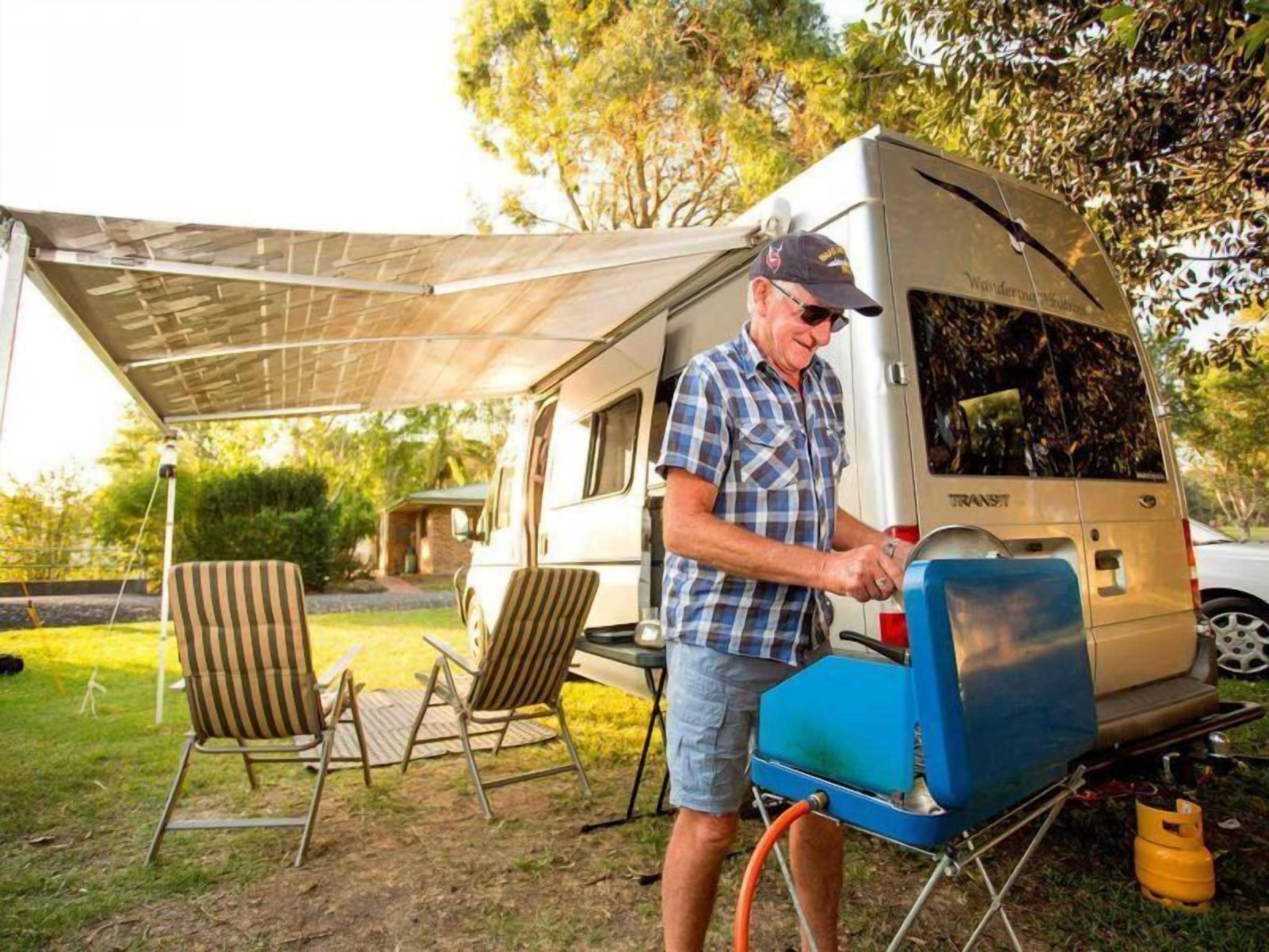 A man in a plaid shirt and cap standing next to a camper van with a blue water heater attached.