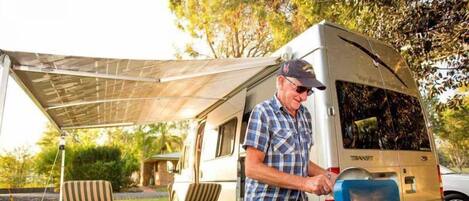 A man in a plaid shirt and cap standing next to a camper van with a blue water heater attached.