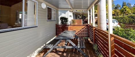 A covered outdoor area with a wooden table and benches, a wooden fence, and a view of a vehicle and greenery in the background.