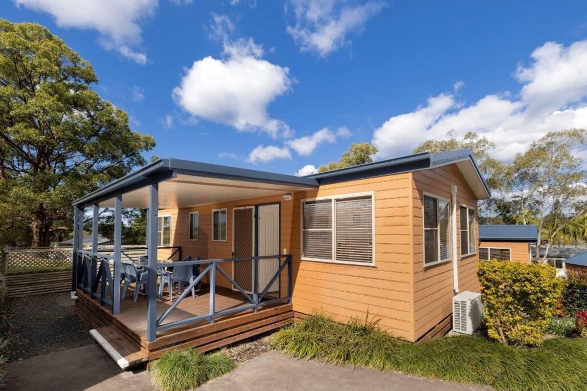 A small, single-story wooden cabin with a covered porch and a small deck, surrounded by trees and a clear sky.