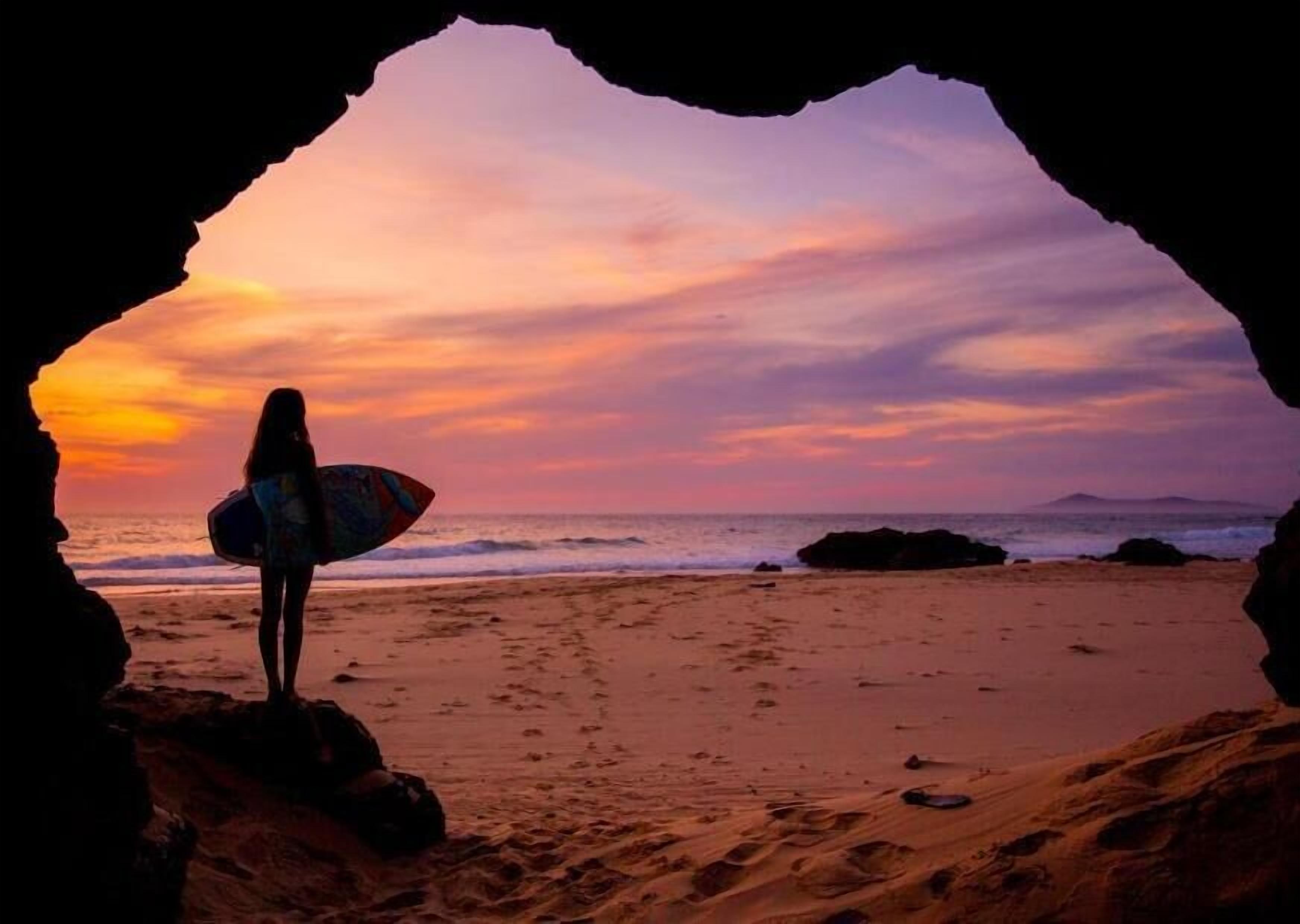 Silueta de una persona sosteniendo una tabla de surf contra un fondo de atardecer, vista a través de la entrada de una cueva.