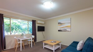 A room with a dining table, chairs, a blue sofa, a coffee table, a TV mounted on the wall, and a window with curtains. - Villa Nova (South Toowoomba)