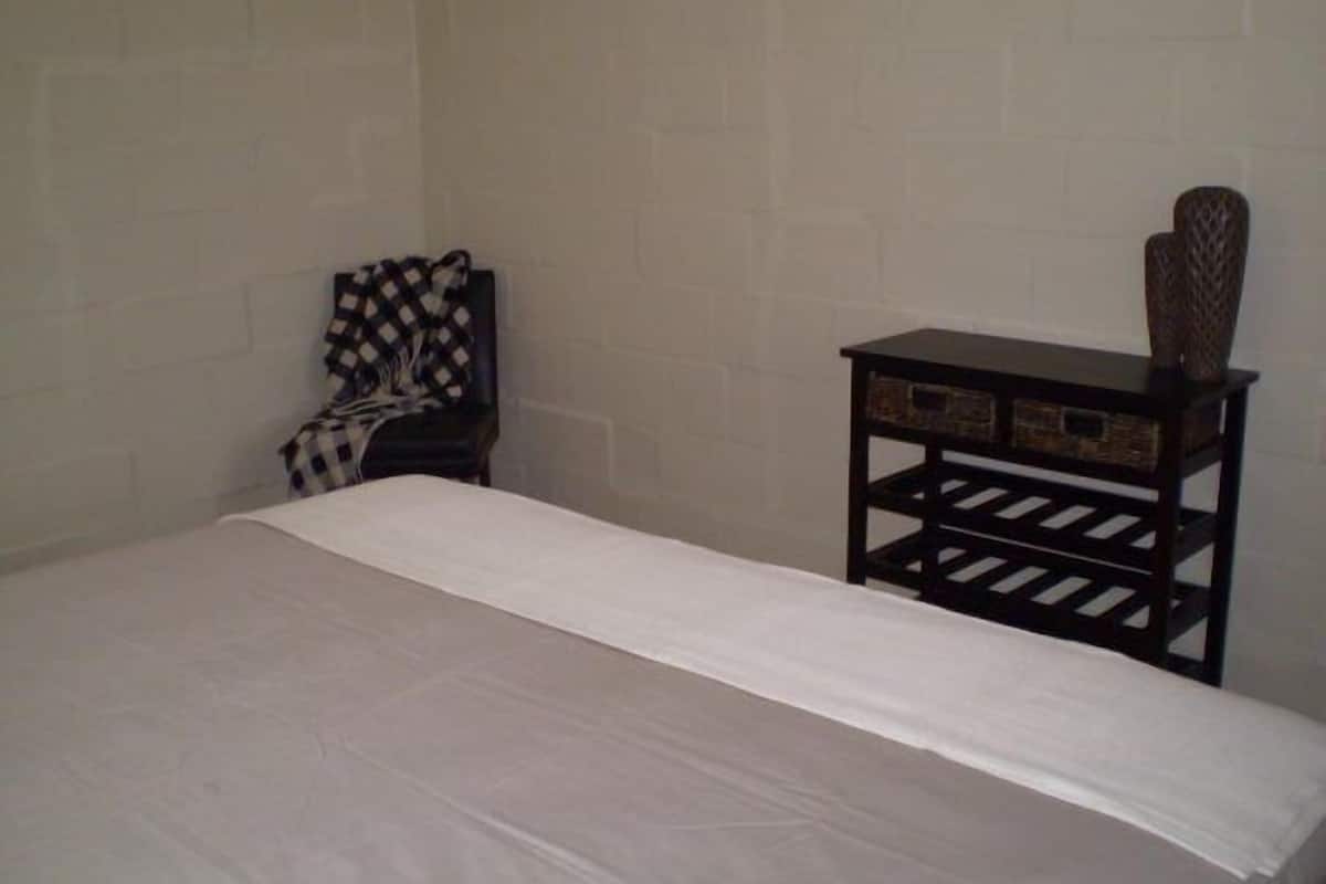 A small, sparsely furnished room with a single bed, a chair with a checkered blanket, and a small table with a decorative vase.