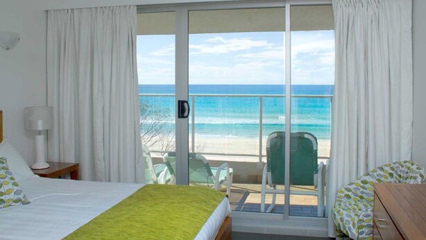 Habitación de hotel con cama, una silla, una mesa y vistas a la playa y al océano a través de amplios ventanales correderas.