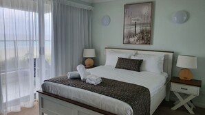Una cama bien hecha con una colcha estampada, dos lámparas de noche, una obra enmarcada en la pared y cortinas translúcidas en la ventana.