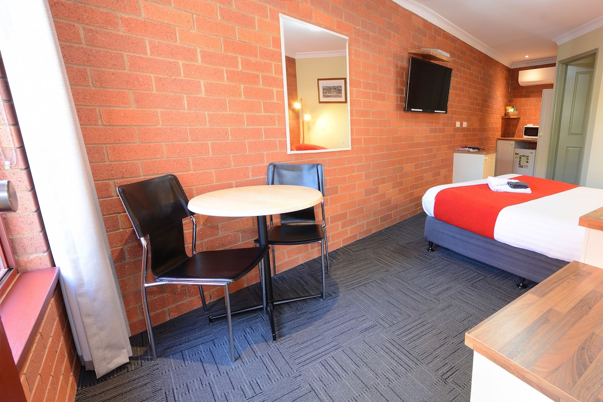 Una habitación de hotel con pared de ladrillo, una cama con colcha rojo y blanco, una mesita con dos sillas, un televisor montado en la pared y un escritorio de madera con una lámpara.