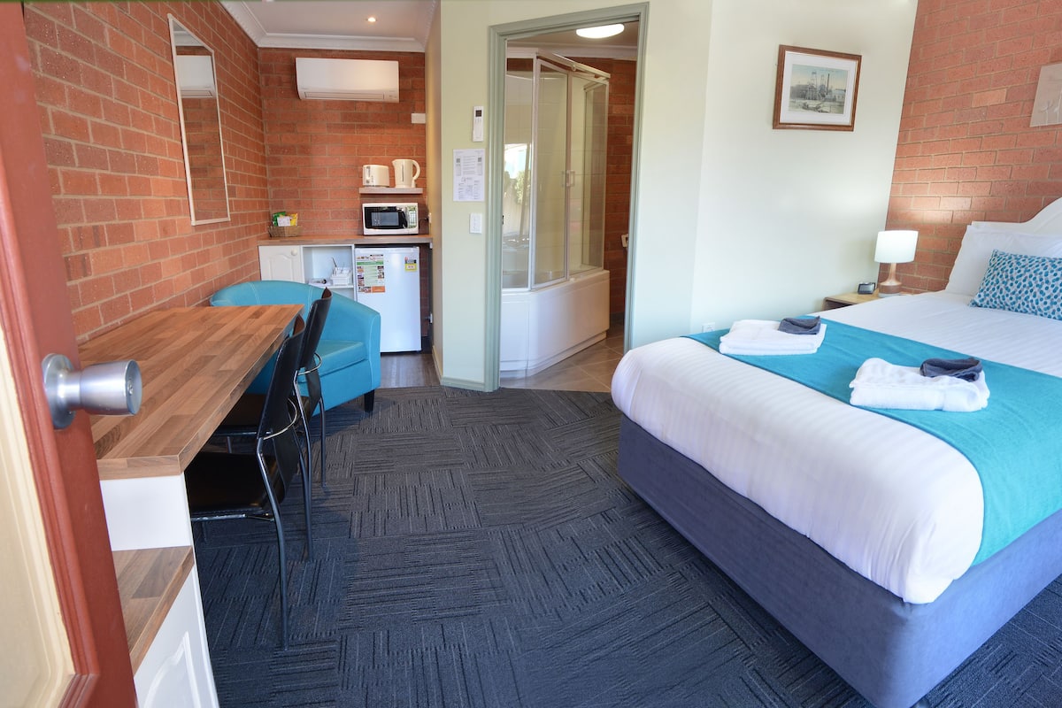 Habitación de hotel con cama, escritorio, silla, microondas y zona de ducha.