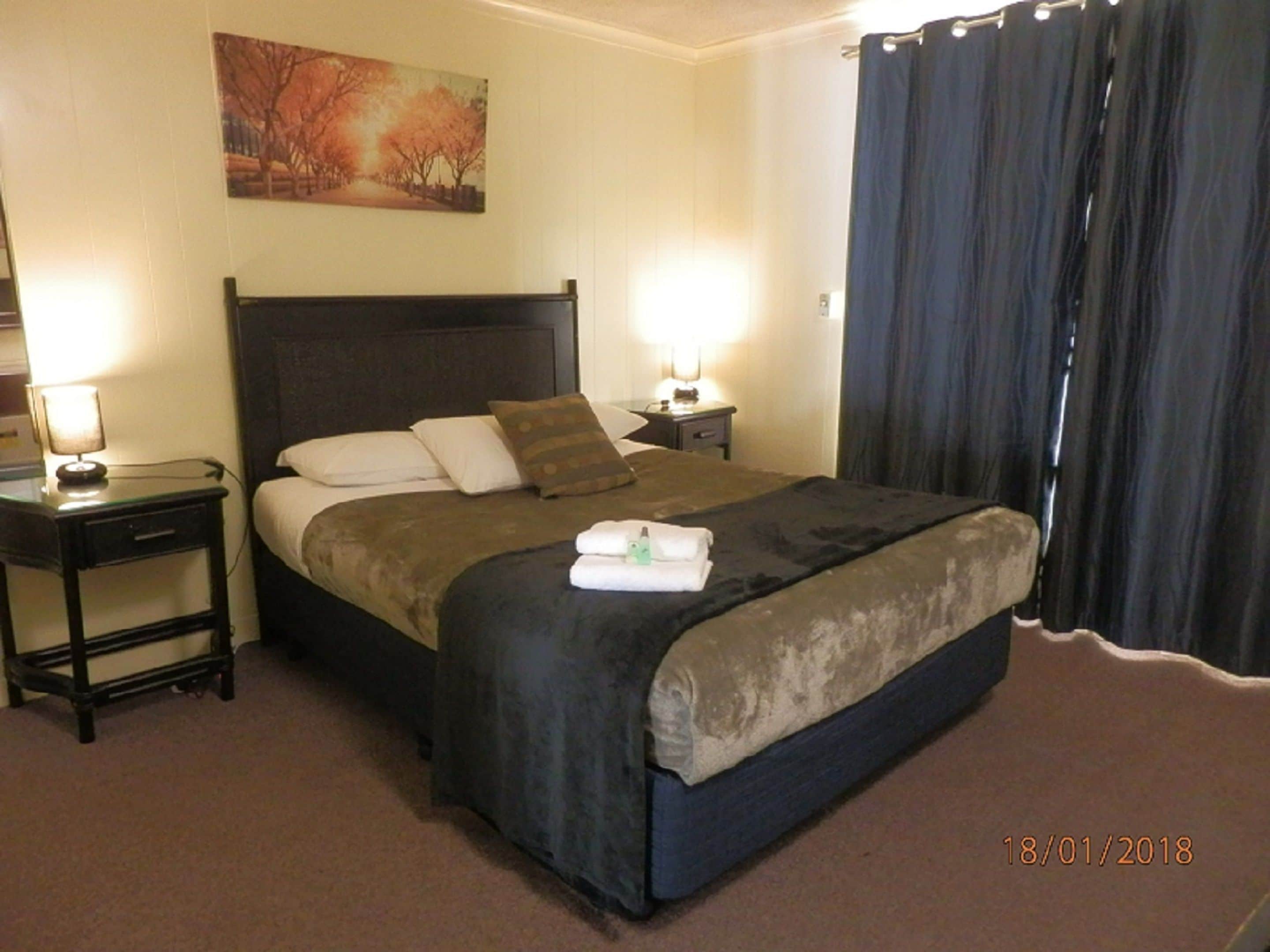 A hotel room with a large bed, two bedside tables with lamps, a painting on the wall, and a dark curtain on the window.