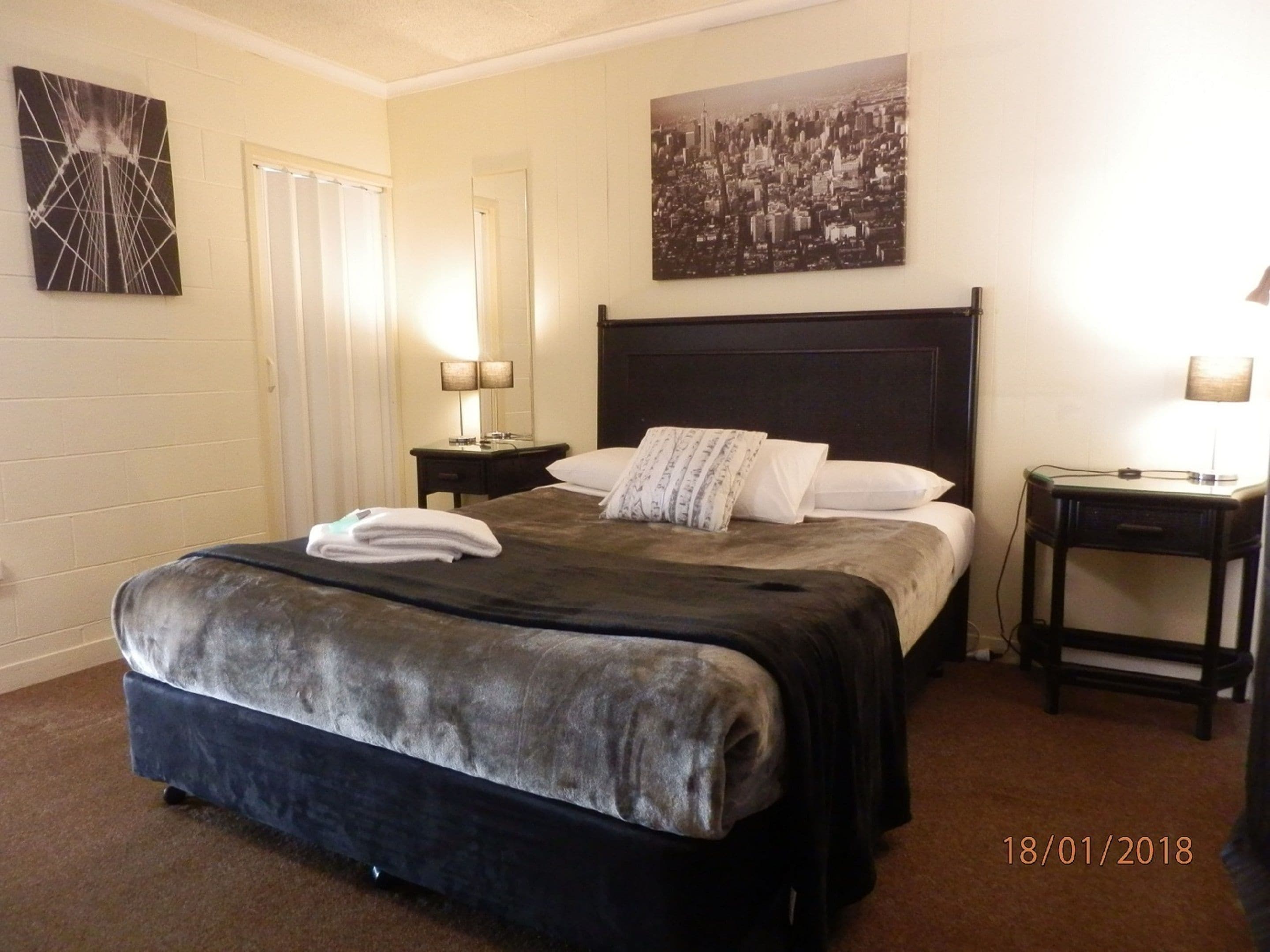 A hotel room with a large bed, a nightstand, a lamp, and a cityscape painting on the wall.