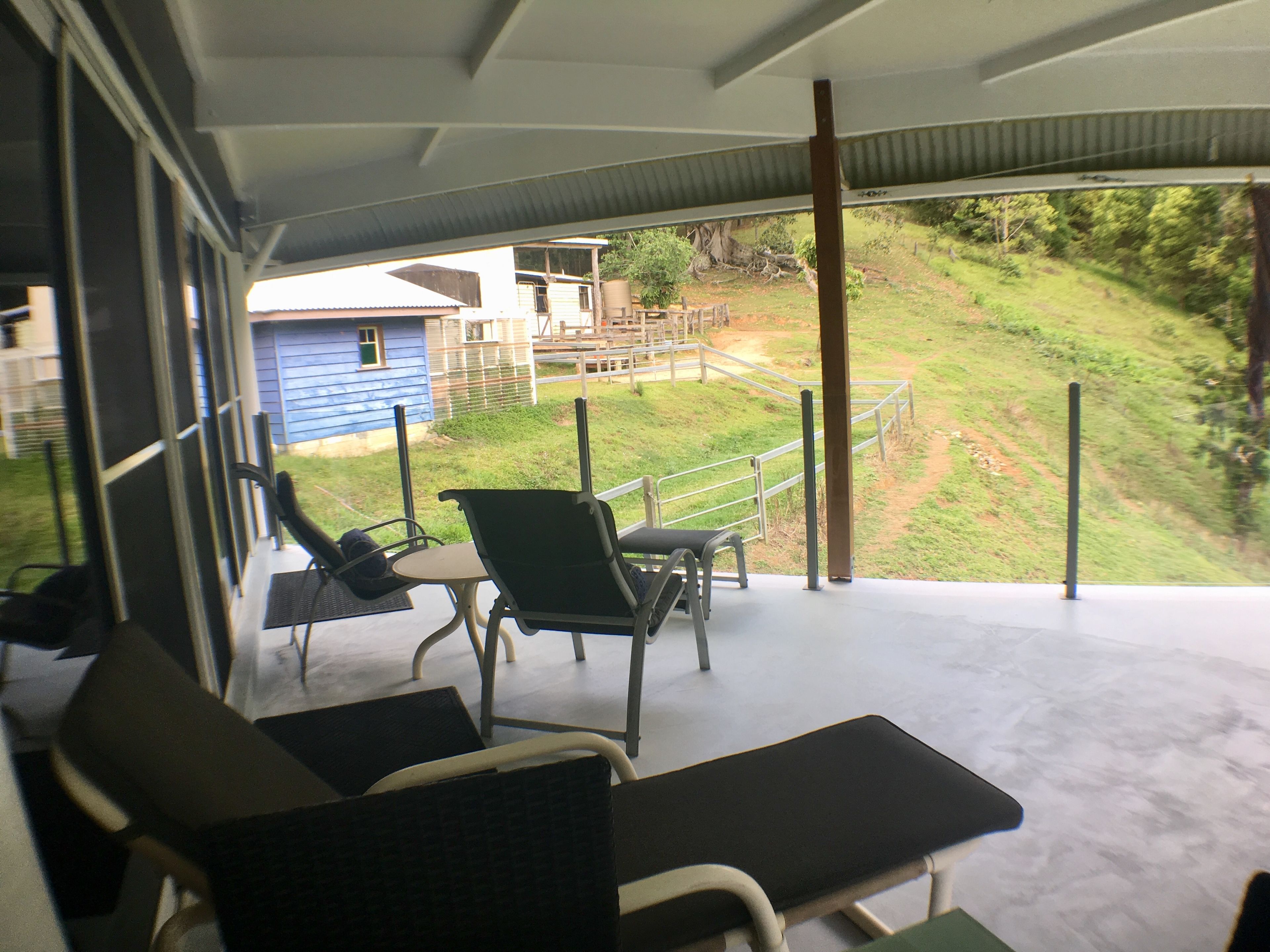 Un espace extérieur abrité, aménagé avec deux chaises et une table, qui offre une vue sur une colline verdoyante avec une maison en arrière-plan.