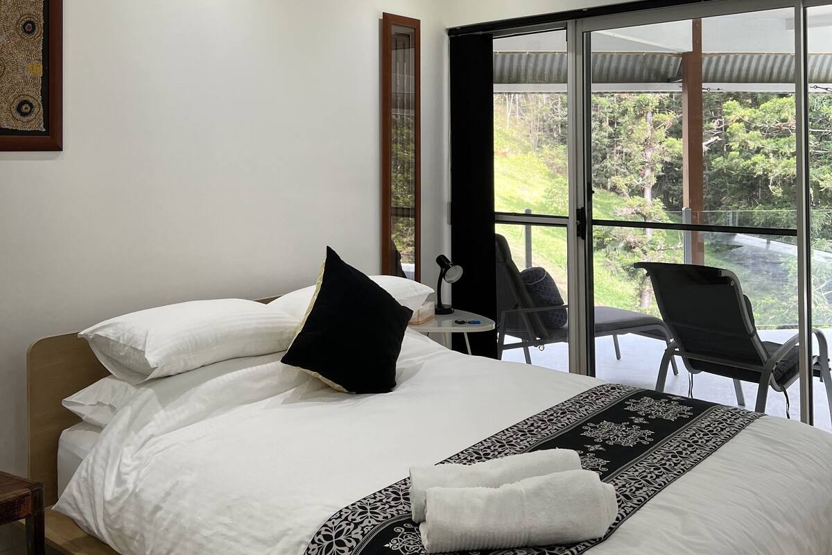 A modern hotel room with a large bed, a ceiling fan, a desk with a chair, and a view of the outdoors through large windows.