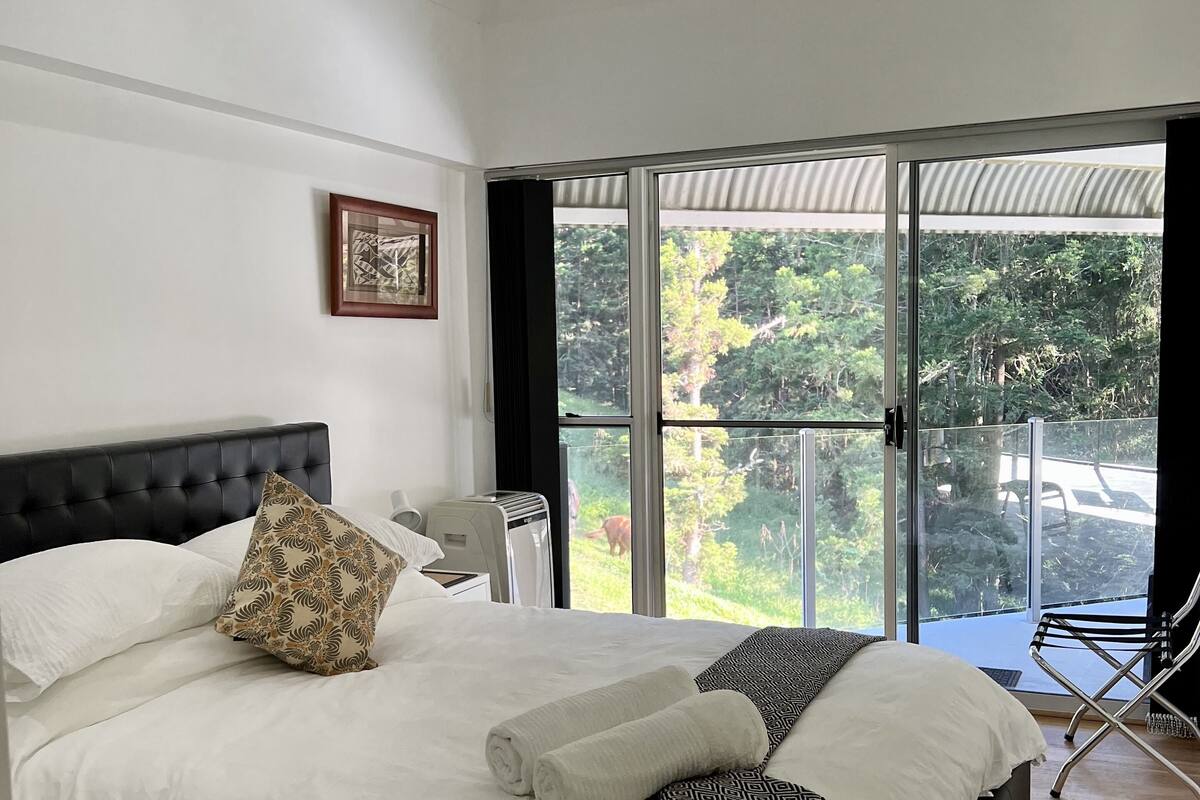 A bedroom with a large bed, a ceiling fan, a window with a view of greenery, and a chair by the window.