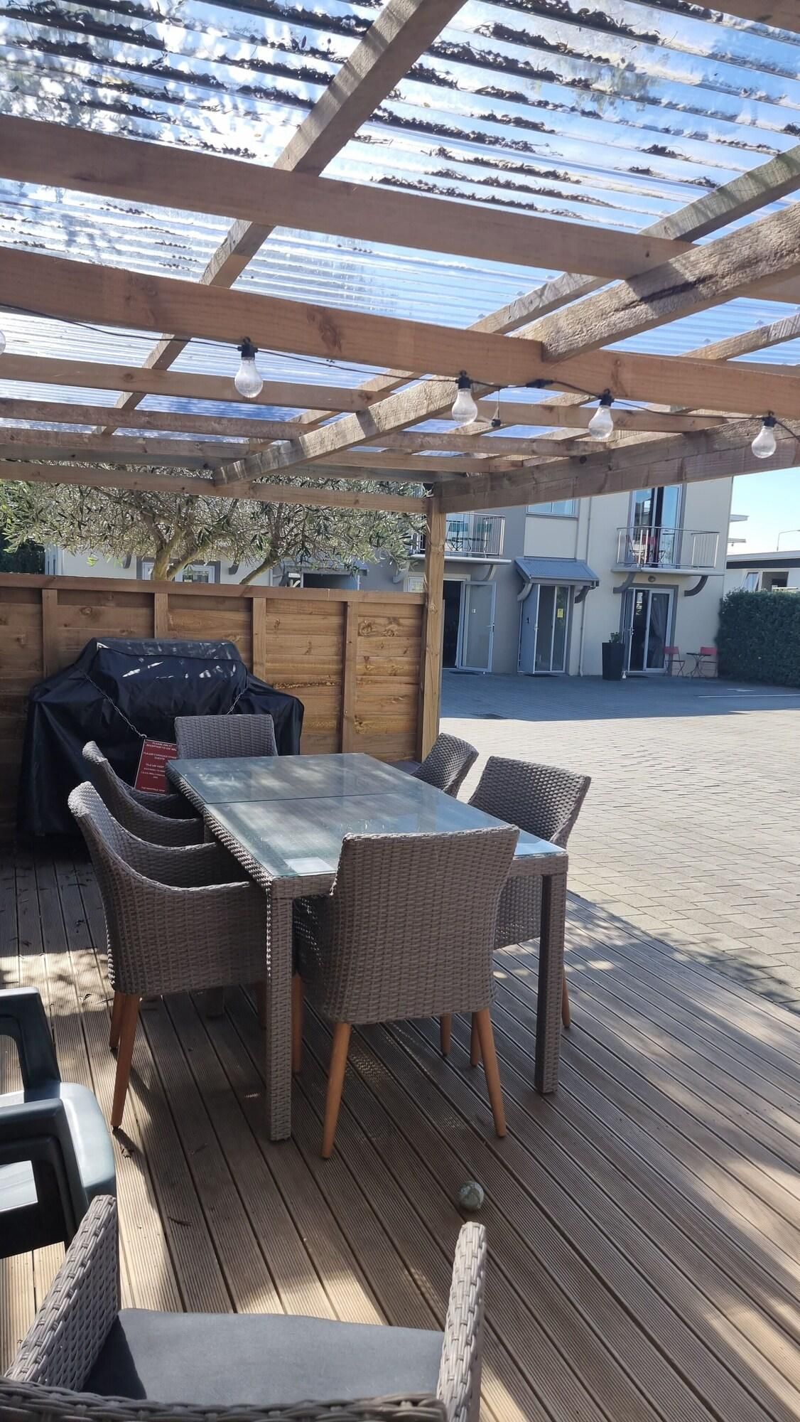 An outdoor patio with a wooden table and wicker chairs, a barbecue grill, and a covered area with a wooden ceiling.