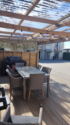 An outdoor patio with a wooden table and wicker chairs, a barbecue grill, and a covered area with a wooden ceiling. - Highfield Mews (Oamaru)