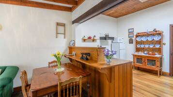 A wooden dining table with chairs, a green sofa, a wooden cabinet with dishes, and a kitchen area with a microwave and shelves.