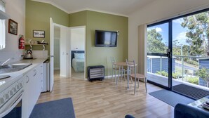 A modern kitchen with a built-in oven and sink, a dining area with a table and chairs, a flat-screen TV mounted on the wall, and a large sliding glass door leading to an outdoor balcony with a view of trees and a road. - Port Huon Cottages (Port Huon)